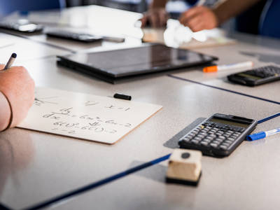Student solving maths calculations on paper with calculator and classroom equipment on desk.