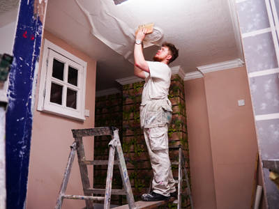 A male student stood on a ladder wallpapering a ceiling 