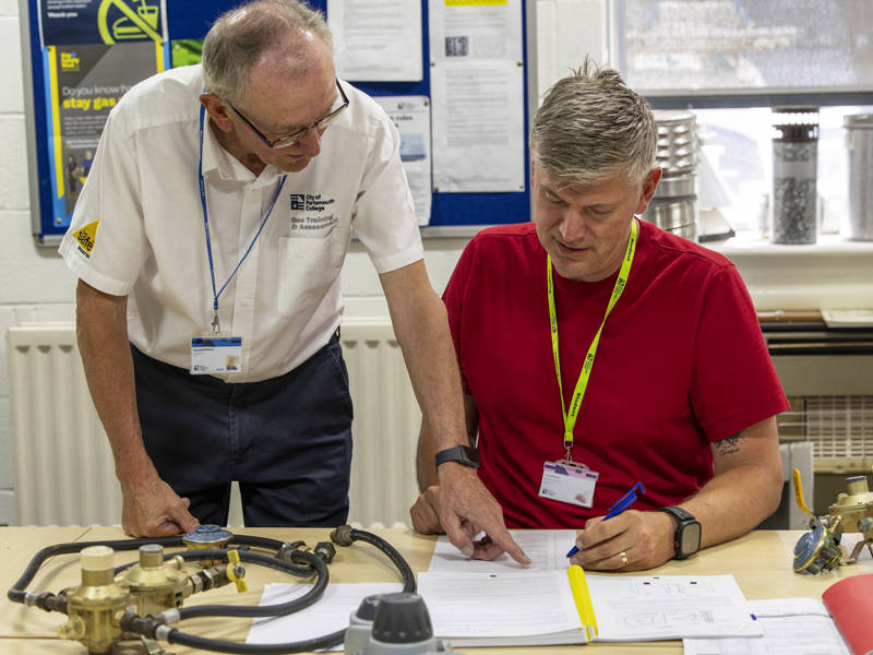 A college tutor supporting an adult learner during a gas training session, reviewing paperwork together at a workbench with gas equipment.