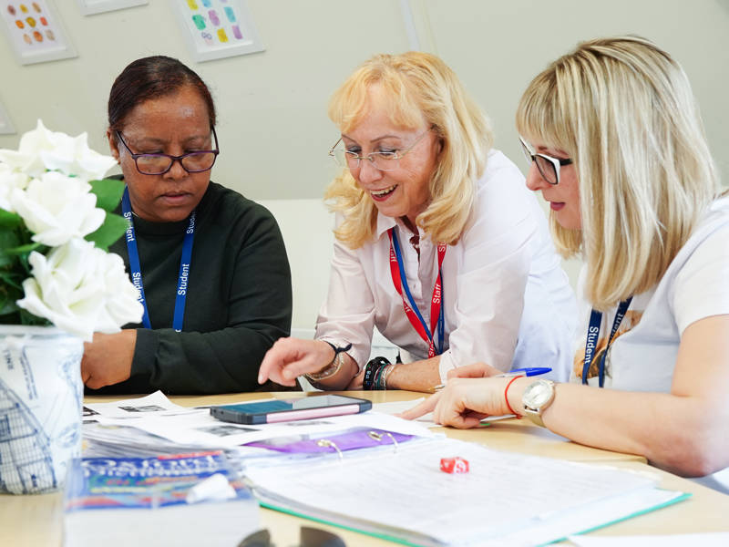 Students sat round staff member working on smart device at desk in classroom.