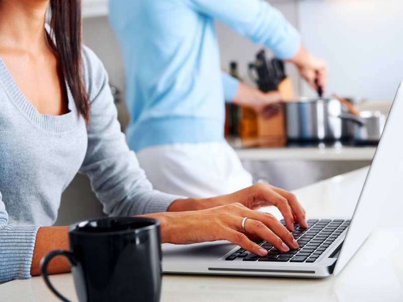 Woman working on laptop in kitchen as partner prepares meal. Happy, healthy relationship. Multiracial couple.